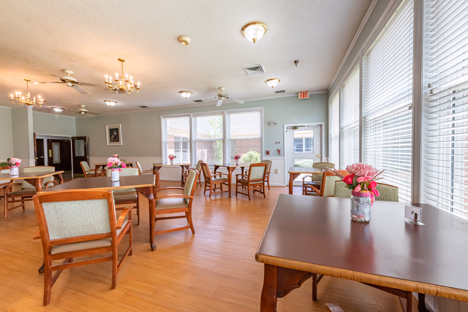 Bright dining room with wooden tables and chairs, floral centerpieces, large windows with blinds, and chandeliers.
