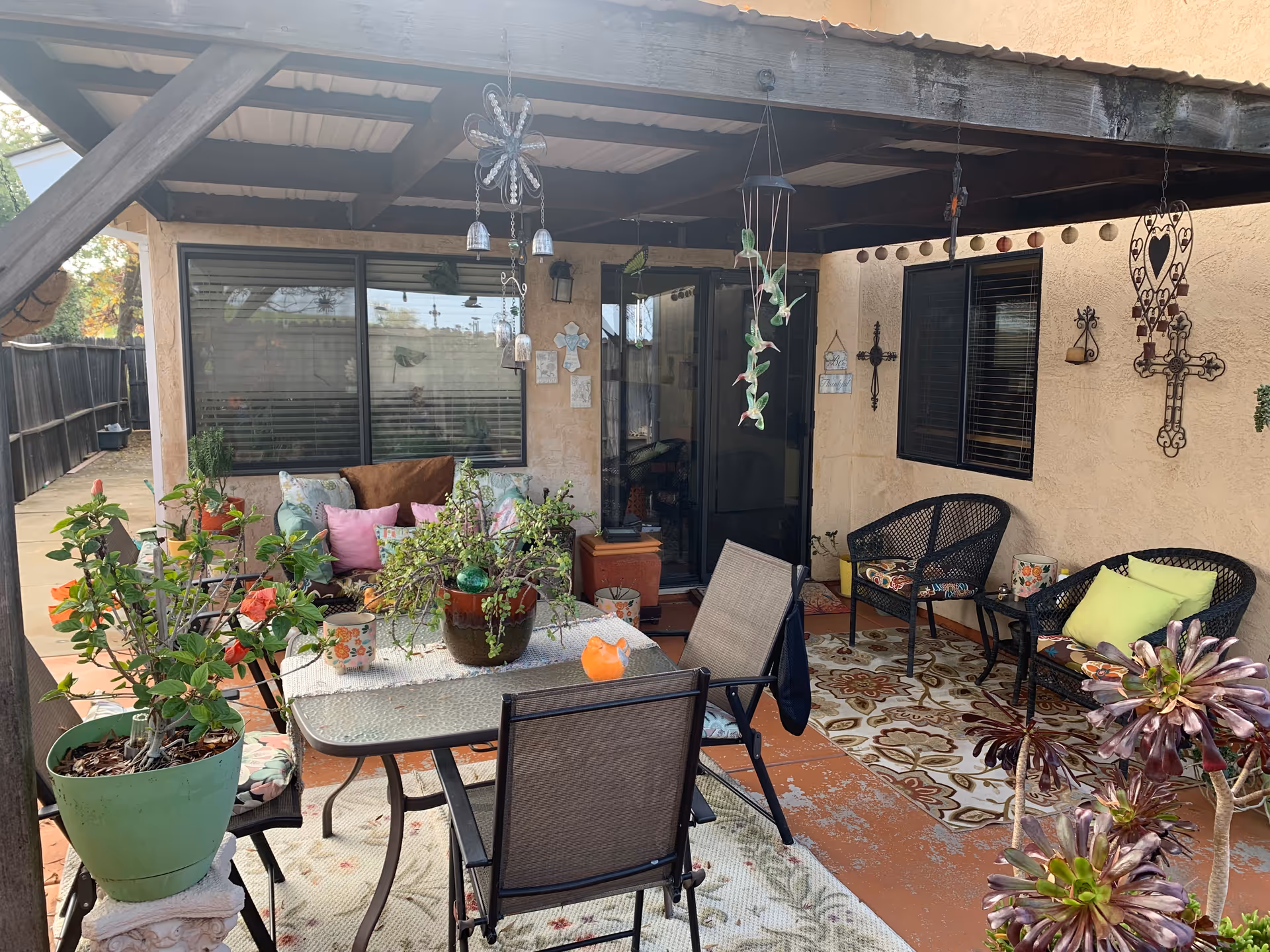 Covered outdoor patio area with a glass table surrounded by chairs, potted plants on the table and around the space, cushioned wicker chairs, decorative hanging ornaments, and wall-mounted crosses on the beige stucco wall.