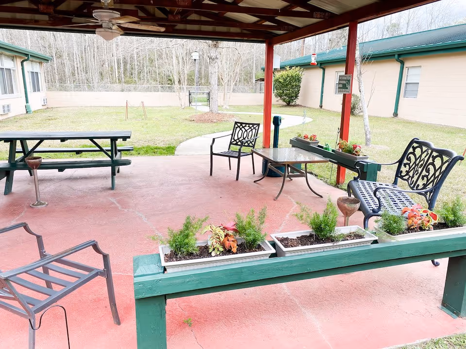 Covered outdoor patio area at Atmore Nursing Center with metal chairs, a glass-top table, a green picnic table, and several flower boxes with plants. The patio overlooks a grassy courtyard enclosed by a beige building and a fence with trees in the background.