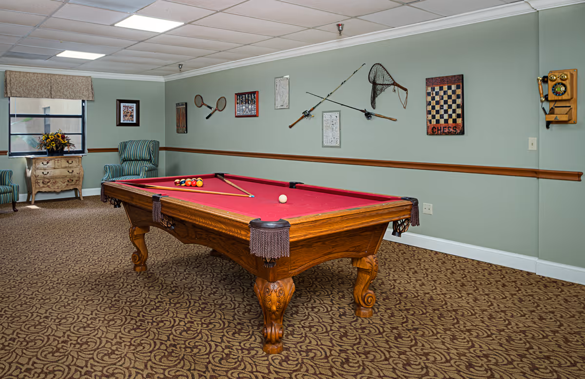 A recreational room with a red felt pool table in the center, two green striped armchairs, a small wooden chest with a flower arrangement, and various wall decorations including fishing rods, a fishing net, tennis rackets, and a chess board. The room has patterned carpet and light green walls.