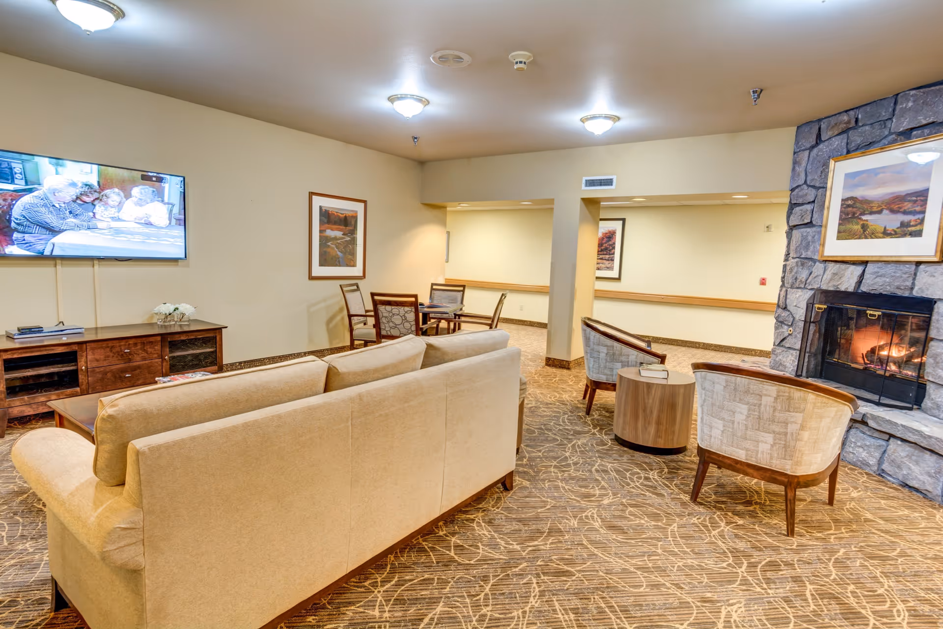 A cozy living room area in The Lodge at Vista View featuring a beige sofa, two upholstered chairs, a stone fireplace with a fire burning, a wooden coffee table, a TV mounted on the wall showing an elderly couple, and framed artwork on the walls. The room has warm lighting and patterned carpet flooring.