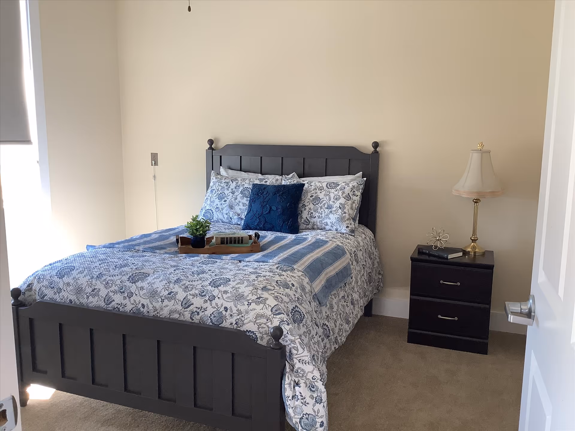 A neatly made bed with a dark wooden frame and floral-patterned bedding in shades of blue and white. There is a small wooden tray with a plant and books on the bed. Next to the bed is a dark wooden nightstand with two drawers, a book, a decorative item, and a lamp with a beige lampshade. The room has beige walls and carpeted floor, with a door partially open on the right side.
