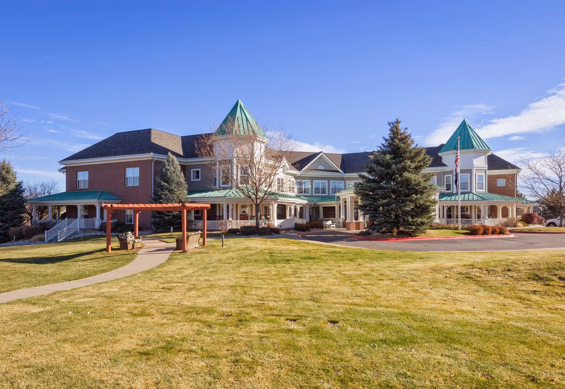 Exterior view of a large senior living facility building with green roofs and brick walls, surrounded by a well-maintained lawn, trees, and a clear blue sky. There is a paved walkway leading to the entrance and a wooden pergola with benches in the foreground.