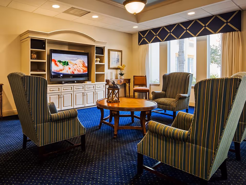 A cozy living room area with four striped armchairs arranged around a round wooden coffee table. A large white entertainment center with a flat-screen TV is against the wall. The room has blue carpet with a subtle pattern, large windows with beige curtains and a decorative valance, and a small wooden side table with a chair and a flower arrangement.
