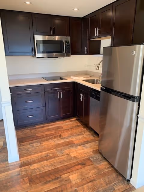 A modern kitchen with dark wood cabinets, stainless steel appliances including a microwave, dishwasher, and refrigerator, light-colored countertops, and wood flooring.