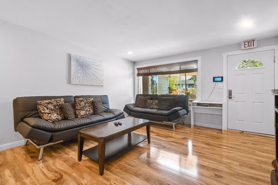 A bright living room with two black leather sofas adorned with patterned cushions, a wooden coffee table with remote controls on it, a large window with wooden blinds, and a white door with an exit sign above it. The room has wooden flooring and white walls with a decorative wall art piece.