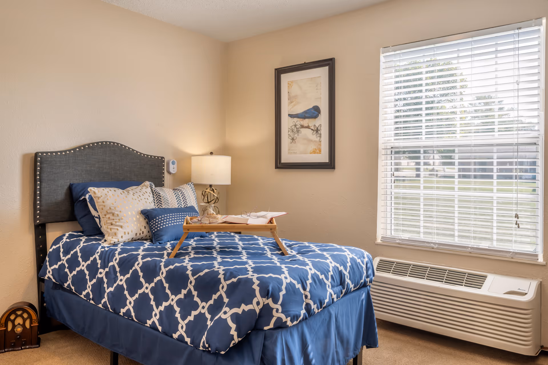 A neatly made single bed with a blue and white patterned comforter and multiple pillows in a small bedroom. A wooden tray with an open book and glasses rests on the bed. There is a bedside table with a lamp and a framed picture of a blue bird on the wall above the bed. A window with white blinds lets in natural light, and an air conditioning unit is installed below the window.