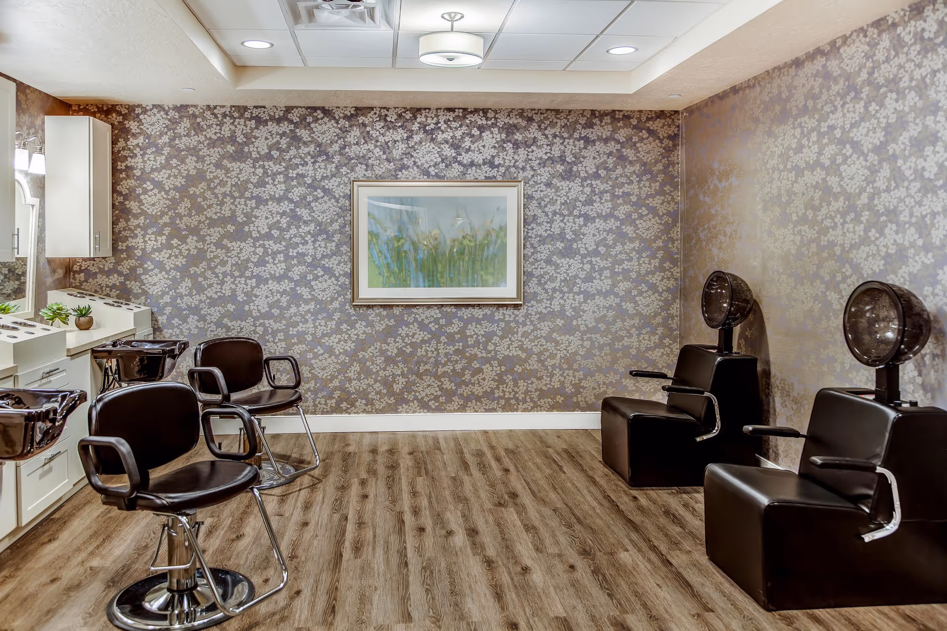 Interior view of a hair salon area with two black salon chairs and two black hair drying chairs against floral patterned wallpaper. There is a framed painting of green plants on the wall and a wooden floor.