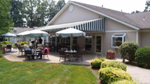 Outdoor patio area at Arden Courts - ProMedica Memory Care Community (Farmington) with tables, chairs, and umbrellas where people are seated, surrounded by greenery and bushes.