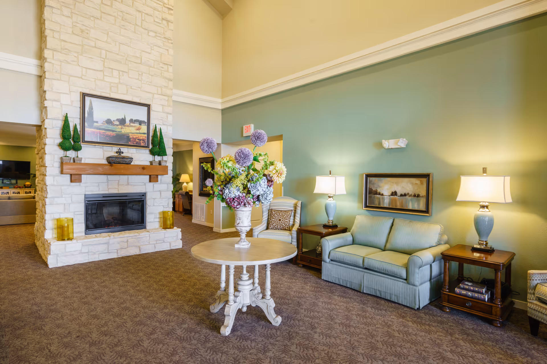 A cozy living room area with a stone fireplace in the center, decorated with small topiary trees and a framed landscape painting above the mantel. In front of the fireplace is a round wooden table with a large floral arrangement. To the right, there is a light blue sofa flanked by two wooden side tables with matching blue lamps. A framed artwork hangs on the green wall behind the sofa. The room has carpeted flooring and soft lighting, creating a warm and inviting atmosphere.