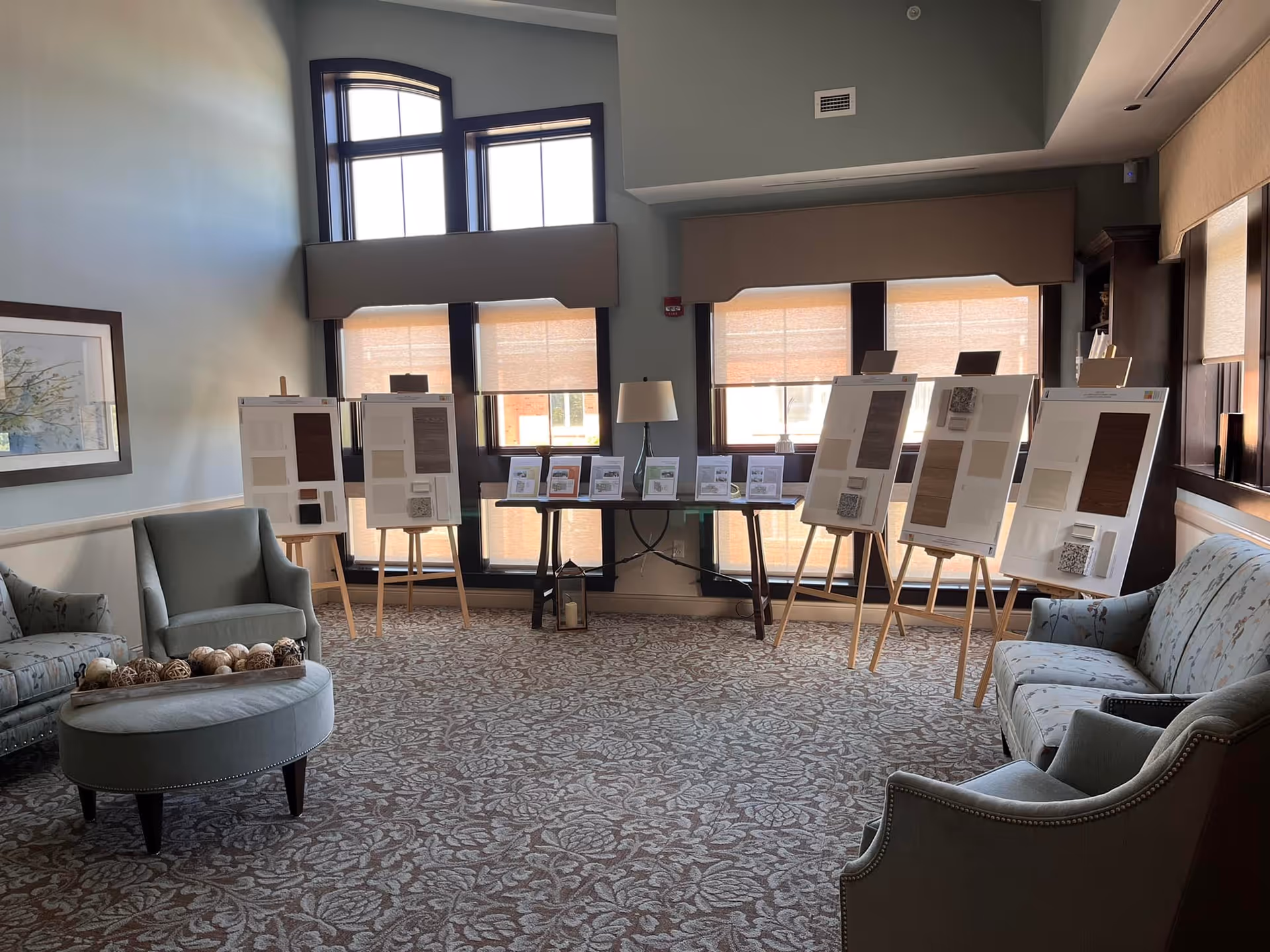 A well-lit room with large windows covered by beige blinds, featuring several easels displaying boards with various samples and information. The room has comfortable upholstered chairs and a round ottoman with decorative balls on it, and a patterned carpet covering the floor.