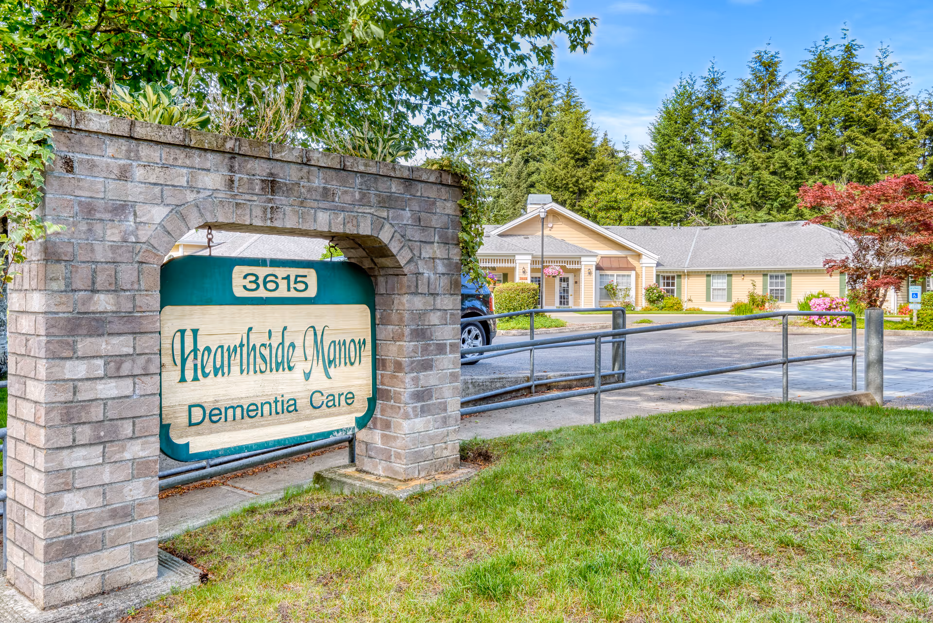 Brick entrance sign reading "Hearthside Manor Dementia Care" with the single-story yellow memory care building and parking area behind it.