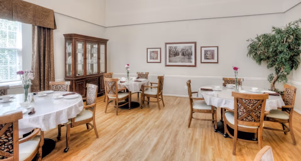 A bright dining room with round tables covered in white tablecloths, each set with plates, cups, and napkins. Wooden chairs with patterned backs surround the tables. A large window with brown curtains lets in natural light. A wooden china cabinet with glass doors displays dishes. Three framed pictures hang on the wall, and a large green potted plant is in the corner.
