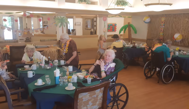 Elderly residents, some in wheelchairs, seated at decorated tables in a communal dining room during a tropical-themed celebration.