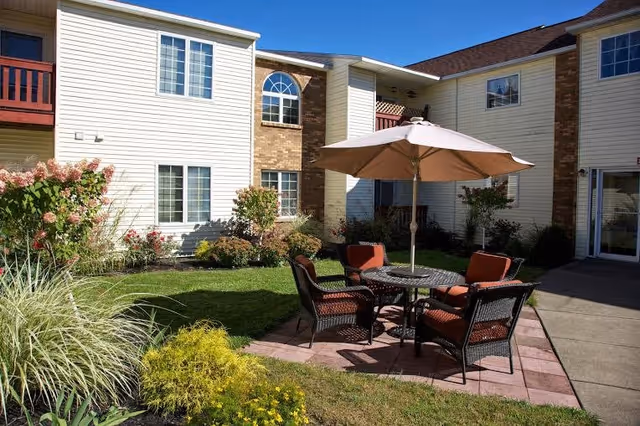 Outdoor patio area with a round table, four cushioned chairs, and a large beige umbrella in the courtyard of a two-story building with white siding and brick accents. The surrounding area has green grass, bushes, and flowering plants under a clear blue sky.