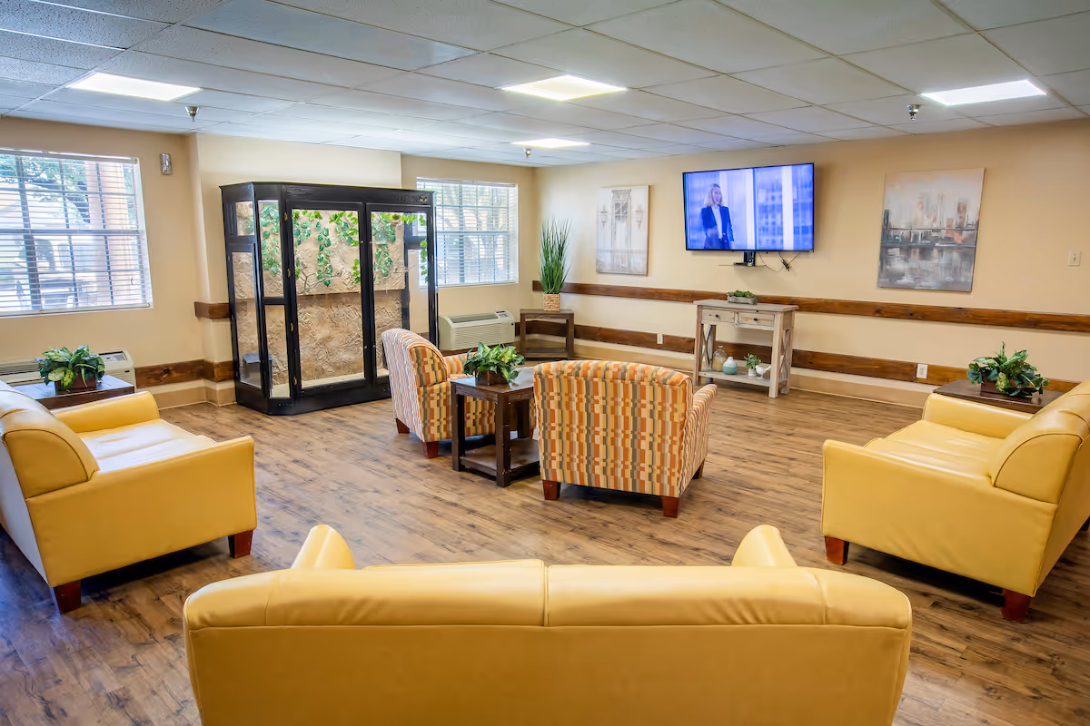 Lounge area in a senior facility with yellow seating, striped chairs, TV, and plants.