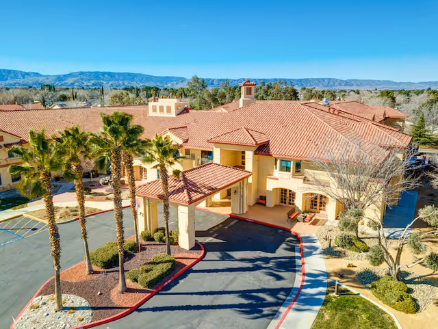 Aerial view of a Mediterranean-style senior living building with red tile roofs, a covered entrance, palm trees, and surrounding landscaping.