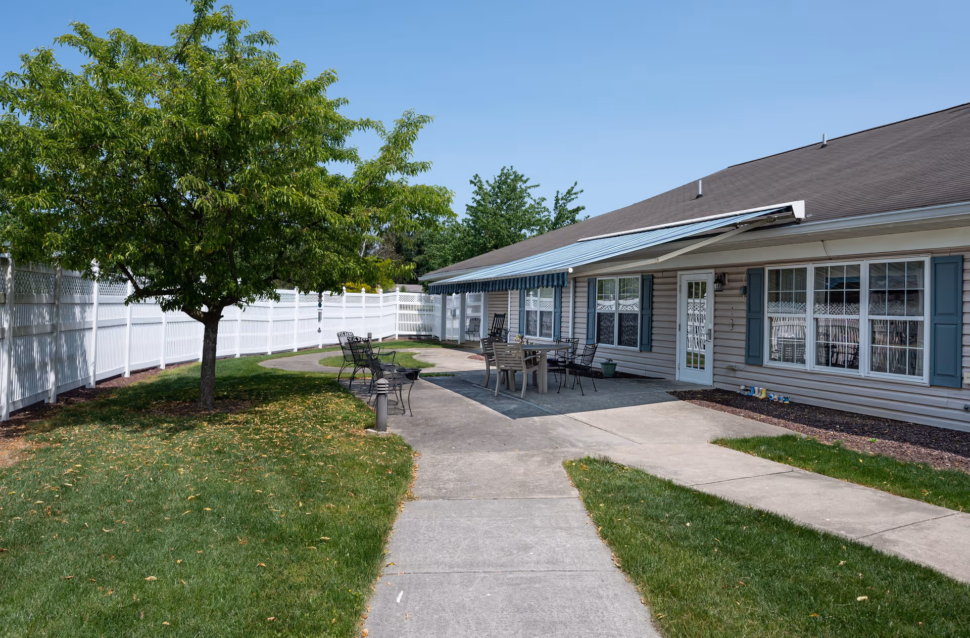 Outdoor patio area of a senior living community with a concrete walkway, green grass, a tree, and a building with windows and a door. The patio has a table with chairs and an awning providing shade.