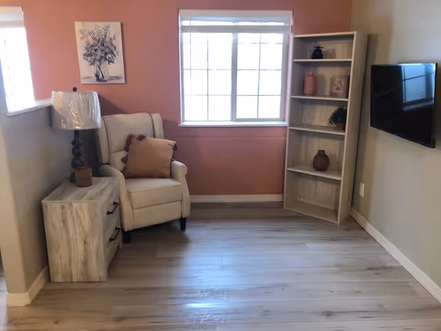 A cozy corner of a living room with light wood flooring, a beige armchair with a brown cushion, a wooden side table with a lamp, a white bookshelf with decorative items, a wall-mounted flat screen TV, and a window with white blinds letting in natural light. The walls are painted in a combination of light beige and muted pink.