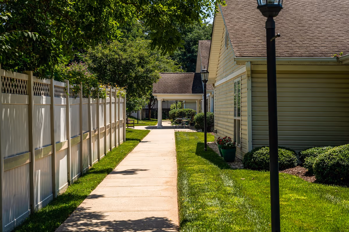 A sunny outdoor walkway bordered by a white fence on the left and a beige building with windows and landscaping on the right. There are green bushes, a potted plant with flowers, and a lamp post along the path. Trees provide shade over part of the walkway, and a covered seating area with a table and chairs is visible in the background.
