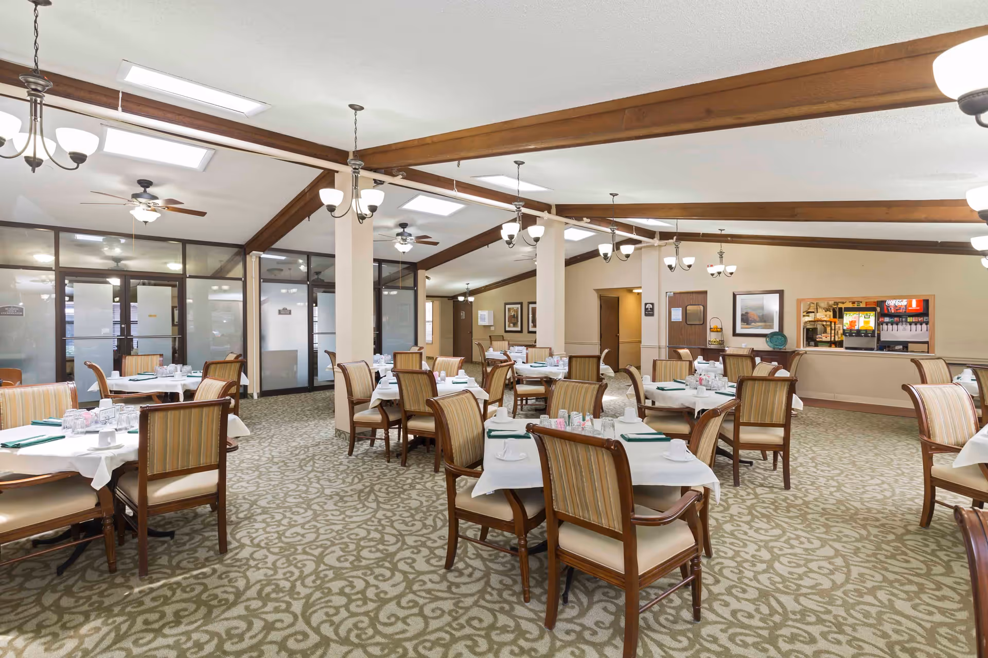 Spacious assisted living dining room with multiple tables set for meals, upholstered chairs, chandeliers, and a beverage station.