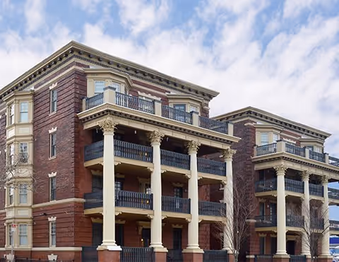 Exterior view of a multi-story brick building with classical architectural elements including large white columns and balconies, under a partly cloudy sky.