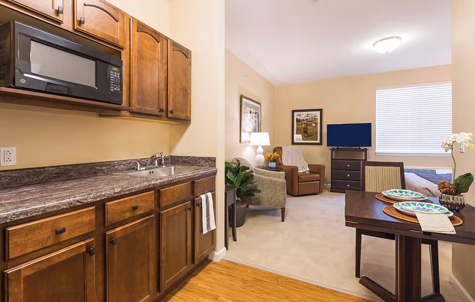 Interior view of a senior living facility apartment showing a kitchenette with wooden cabinets, a microwave, and a sink on the left. The room opens into a living area with a brown armchair, a small table with a lamp, a TV on a dresser, and a dining table set with plates and placemats. A window with blinds is visible in the background.