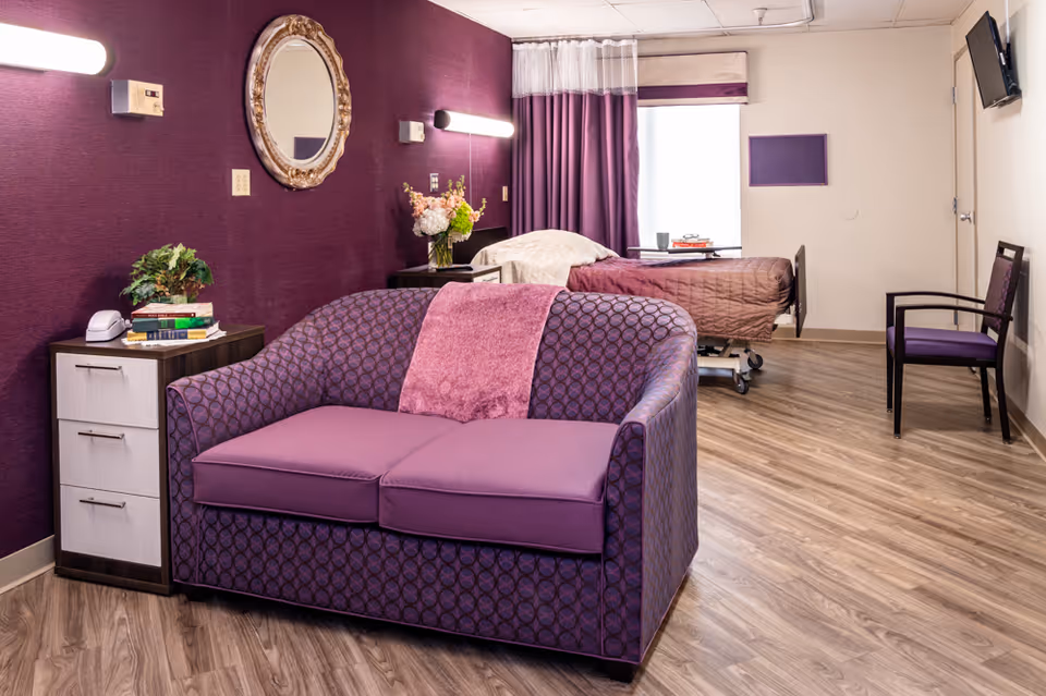 A nursing facility room with a purple patterned loveseat in the foreground, a bedside table with books and a plant, a hospital bed with a brown quilt near a window with purple curtains, a wall-mounted TV, and a chair against the wall. The walls are purple and beige, and the floor has wood-like vinyl flooring.