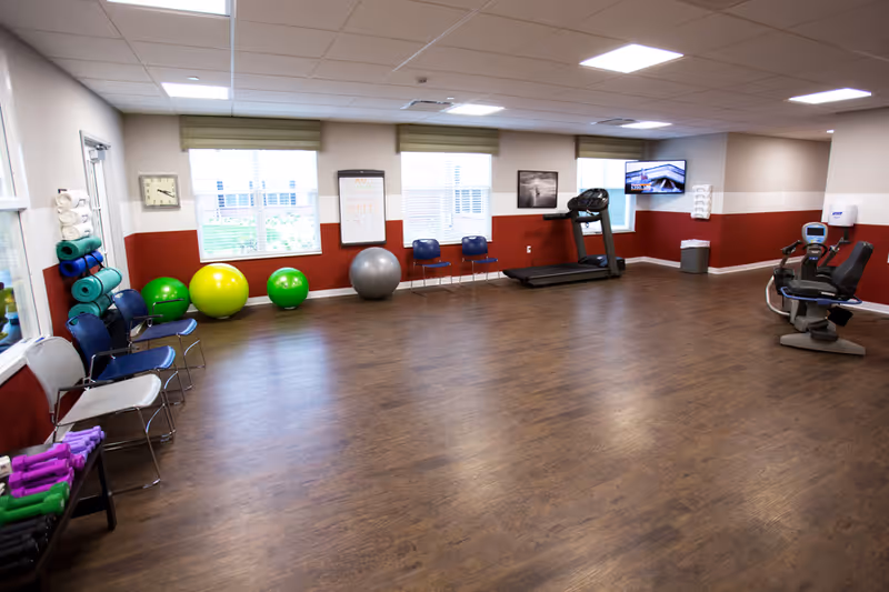 A spacious exercise room with wooden flooring and red and white walls. The room contains exercise balls, yoga mats, dumbbells, chairs, a treadmill, and a recumbent exercise bike. There are windows letting in natural light, a wall clock, a whiteboard, and a mounted TV screen.