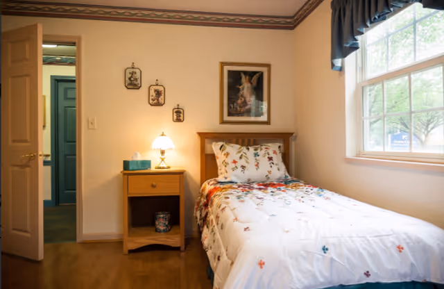 A cozy bedroom with a single bed covered in a white floral comforter and matching pillow. Next to the bed is a wooden nightstand with a lamp and a box of tissues. The room has light-colored walls adorned with small framed pictures and a larger framed artwork above the bed. A window with blue curtains lets in natural light, and an open door reveals a glimpse of a hallway with teal-colored doors.