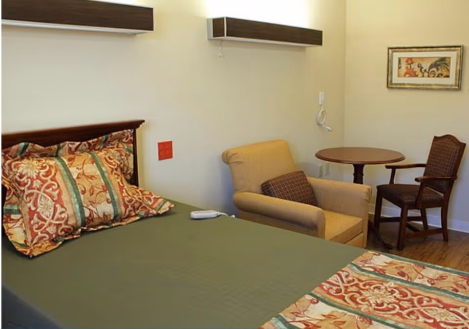 A furnished senior living bedroom with a bed and patterned pillows, an armchair, small round table with a chair, and wall art.