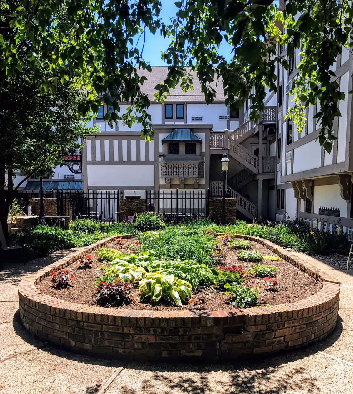 A landscaped garden area with a raised brick flower bed containing various green plants and flowers, surrounded by a paved walkway. The garden is located in the courtyard of a multi-story building with white walls and brown trim, featuring outdoor staircases and windows. Tree branches with green leaves hang overhead, partially shading the area.
