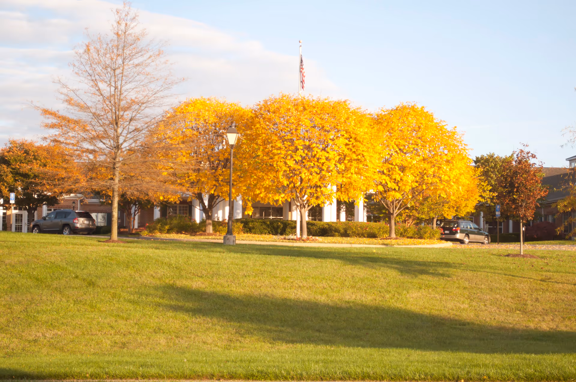 A senior living facility building partially visible behind a row of trees with bright yellow autumn leaves. There is a green grassy lawn in the foreground, a lamppost, and a flagpole with an American flag. Several parked cars are visible near the building.