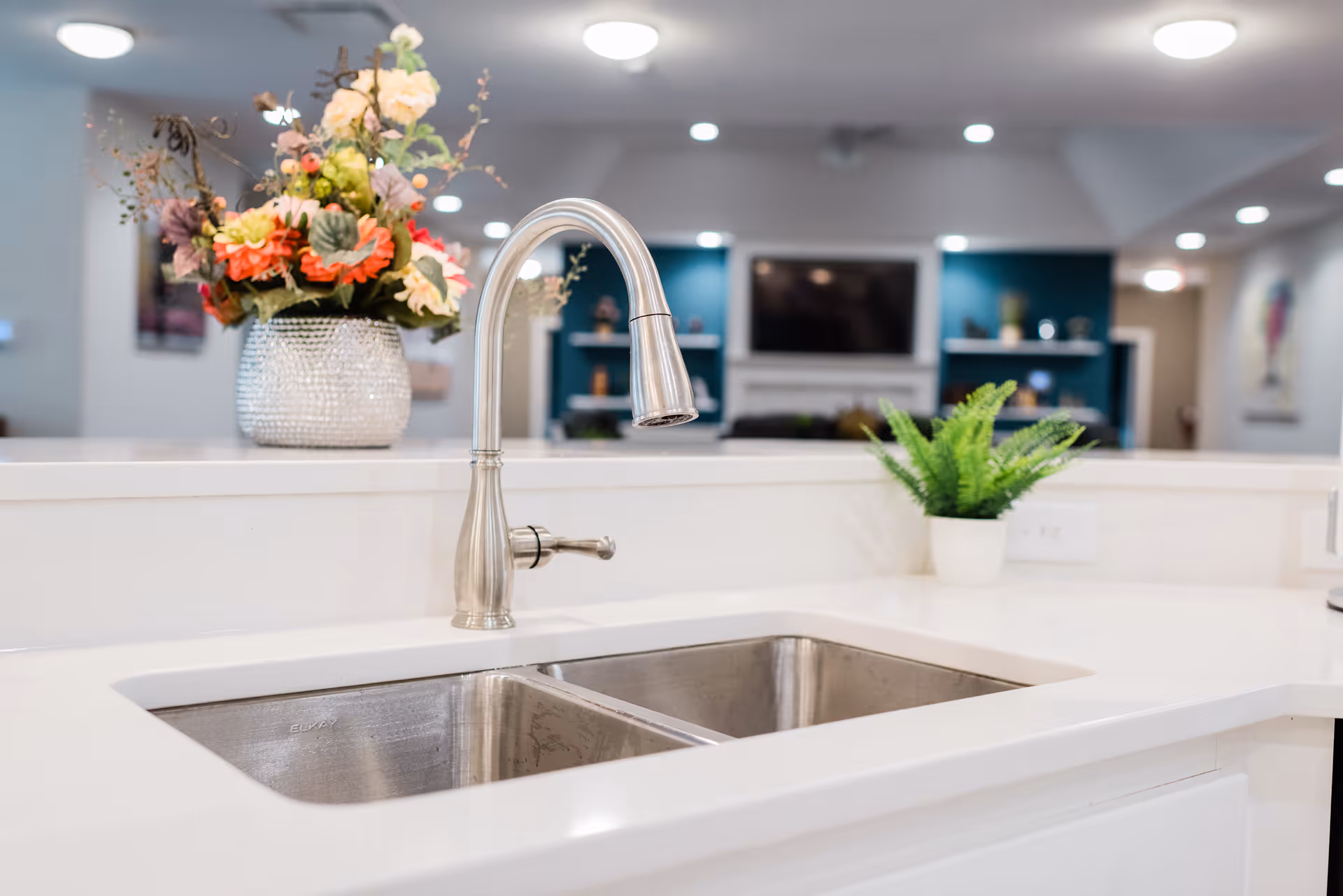 Modern kitchen sink with a stainless steel faucet on a white countertop, decorated with a vase of colorful flowers and a small green potted plant, with a blurred living room area in the background featuring a TV and shelves.