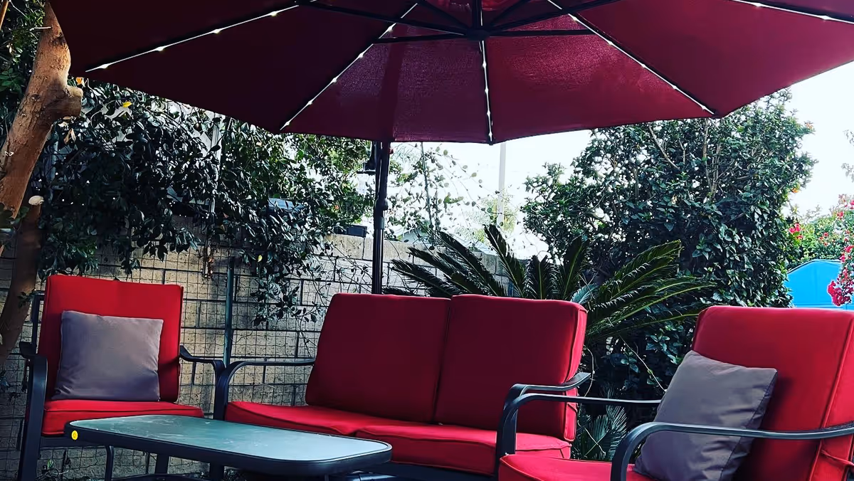 Outdoor seating area with red cushioned chairs and a sofa around a black table, shaded by a large red umbrella. The background includes green plants and a tree.