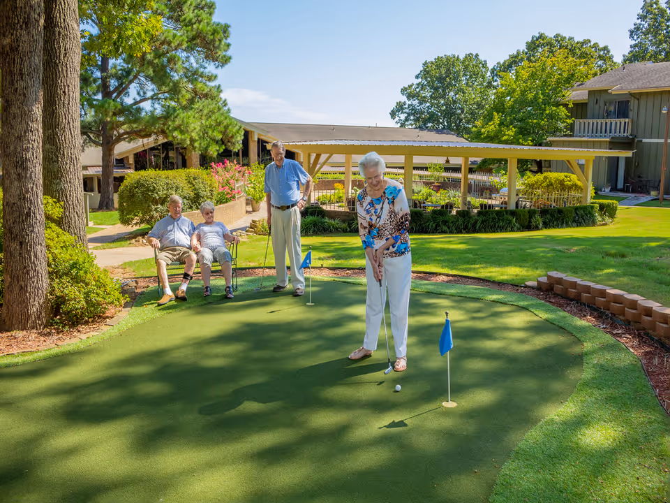 Four elderly people enjoying a sunny day on a putting green at a senior living facility. One woman is putting a golf ball while two people sit on a bench and one man stands nearby watching. The background shows trees, bushes, and buildings under a clear blue sky.