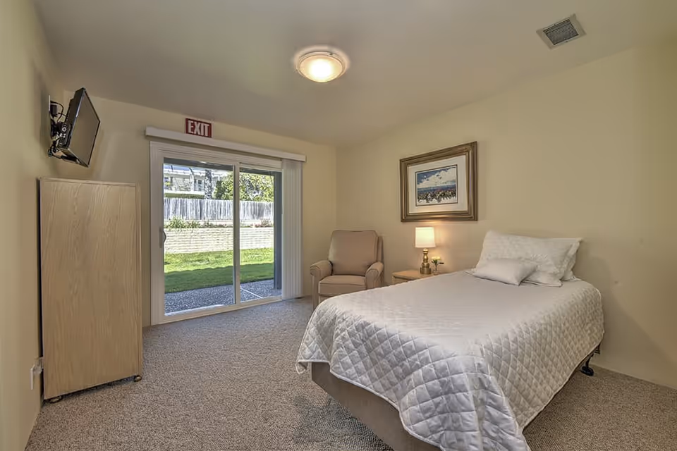A simple bedroom with a single bed covered in a white quilt, a beige armchair, a wooden nightstand with a lamp, a framed picture on the wall, a wall-mounted TV, and sliding glass doors leading to a grassy outdoor area.