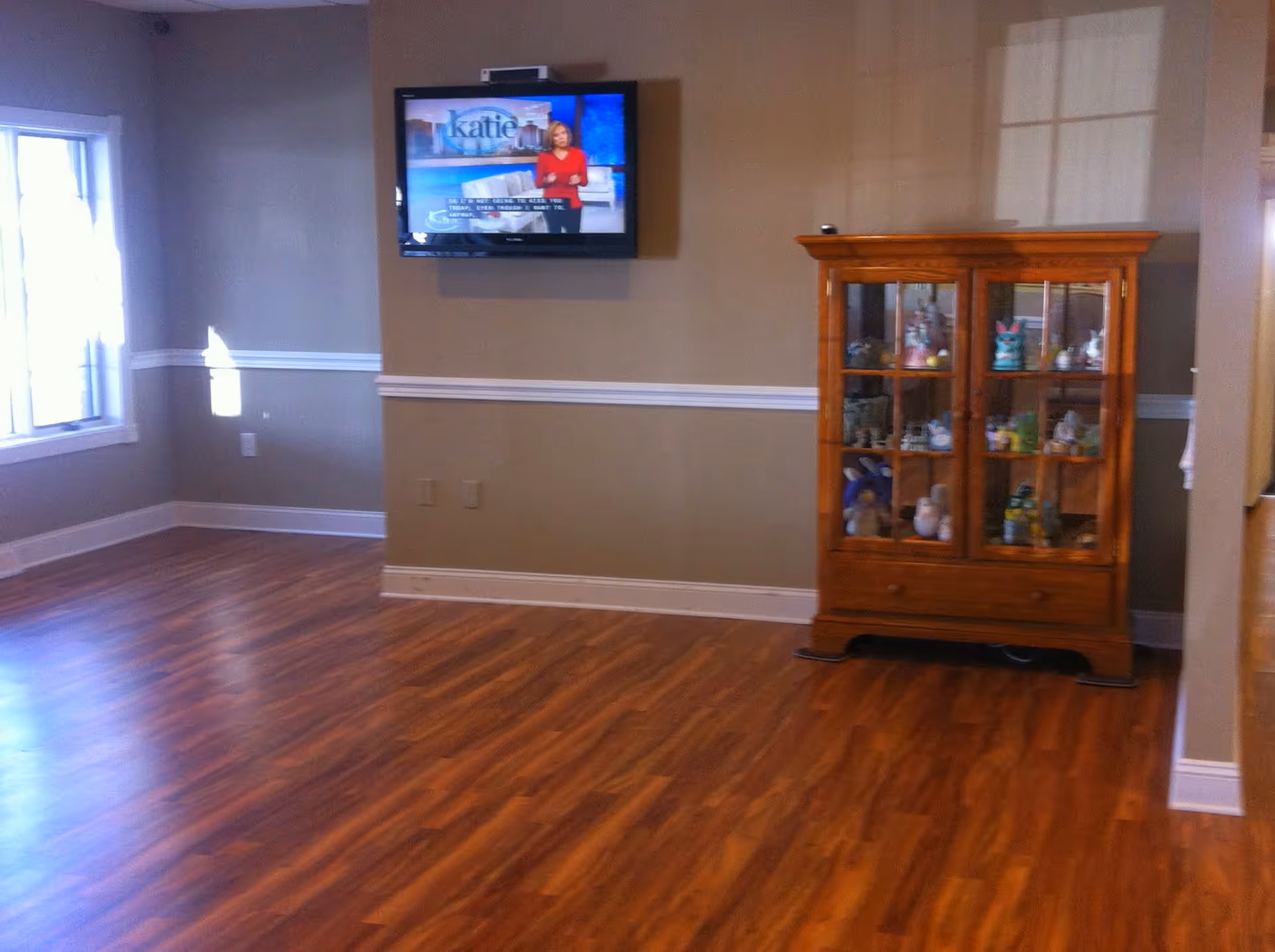 A room with wooden flooring, beige walls, a window on the left letting in natural light, a wall-mounted flat-screen TV displaying a news program, and a wooden glass-front cabinet containing various decorative items.