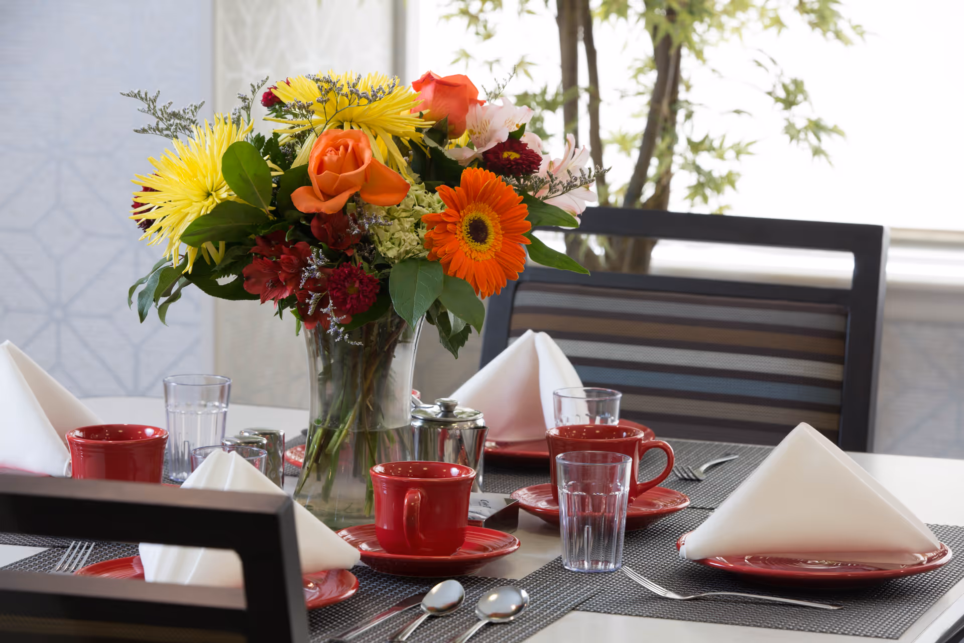 A dining table set with red cups and saucers, clear drinking glasses, silverware, and white folded napkins. A vase with a colorful bouquet of flowers including orange roses, yellow daisies, and other mixed flowers is placed in the center of the table. The background shows a window with natural light and a plant.