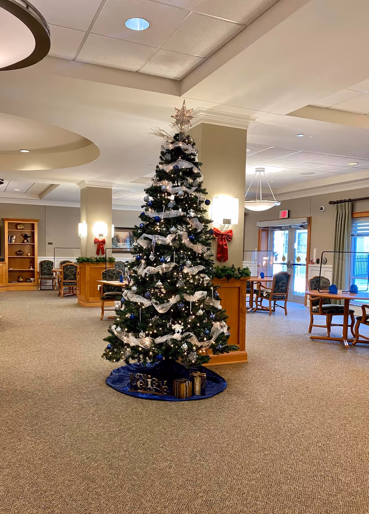 A decorated Christmas tree with white ribbon, blue and silver ornaments, and lights stands in the center of a spacious, carpeted common area with tables, chairs, and festive red bows on the walls. The room has beige walls, ceiling lights, and large windows letting in natural light.