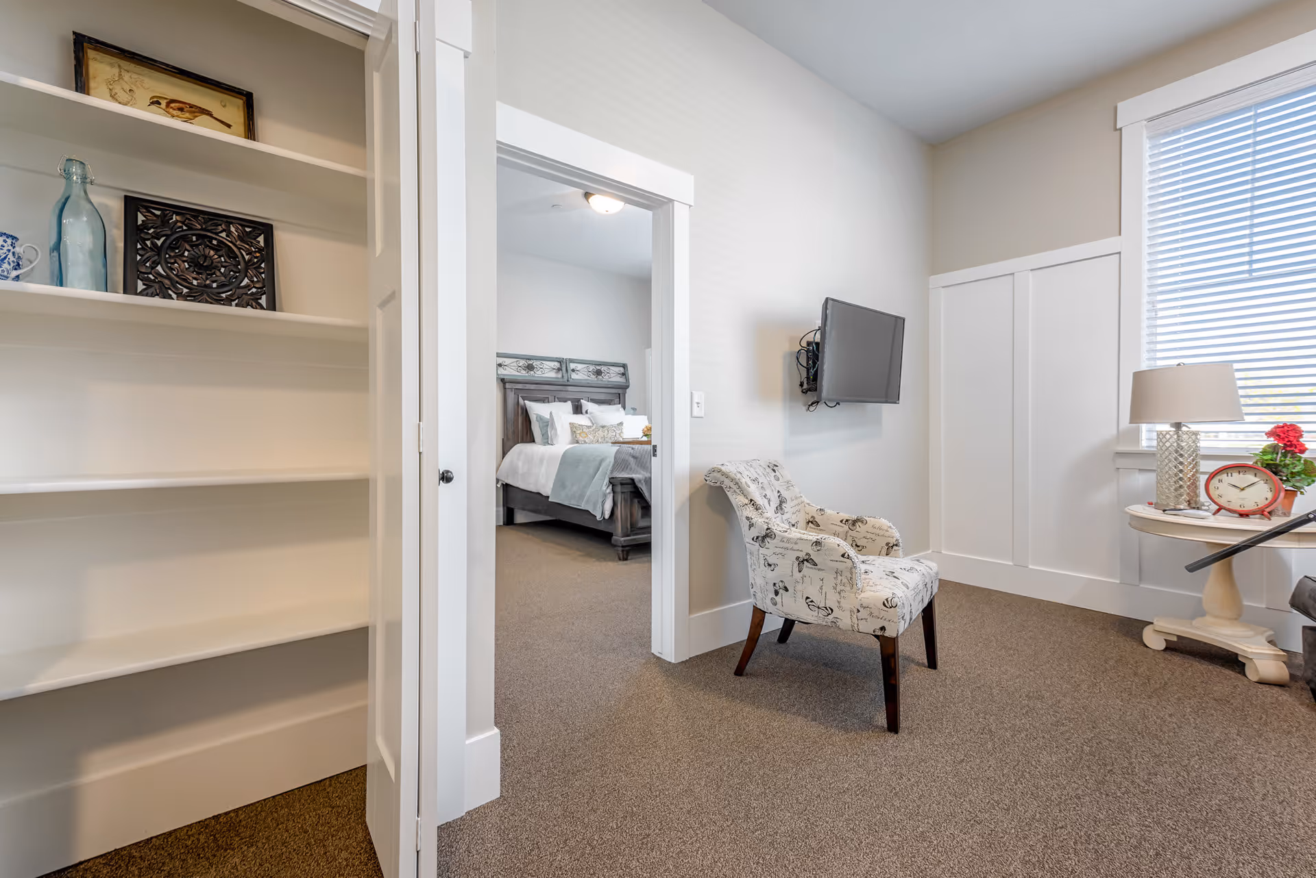 A cozy interior space featuring a patterned armchair facing a wall-mounted flat screen TV. To the right, there is a round side table with a lamp, a red clock, and a small plant near a window with blinds. On the left, an open closet with shelves displays decorative items including a framed bird picture and a blue bottle. Through an open doorway, a bedroom with a wooden bed frame, white bedding, and pillows is visible.
