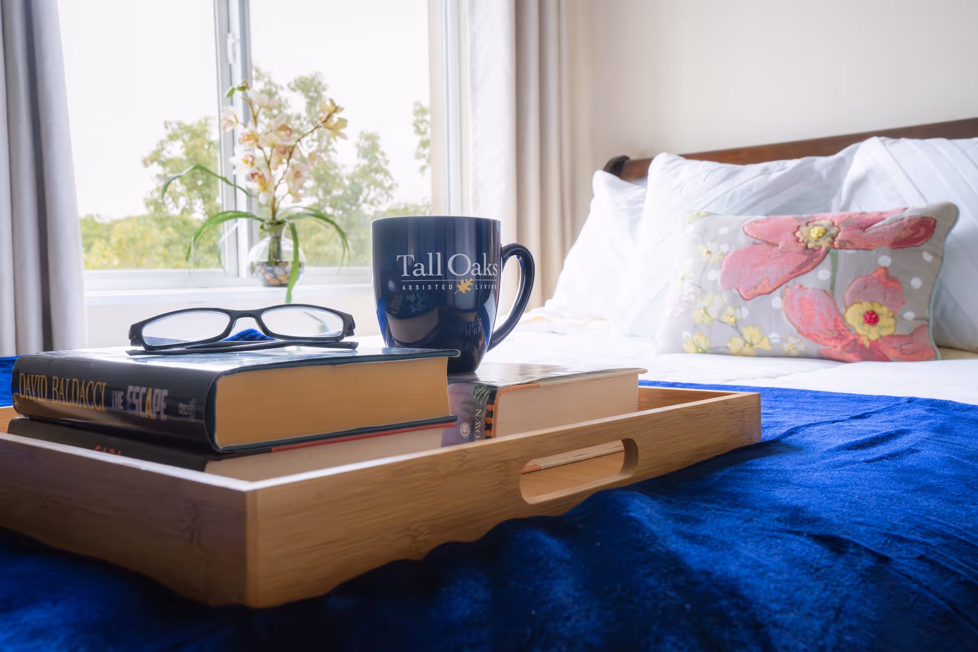 A sunlit bedroom with a bed, decorative pillows and a wooden tray holding books, glasses, and a Tall Oaks mug.