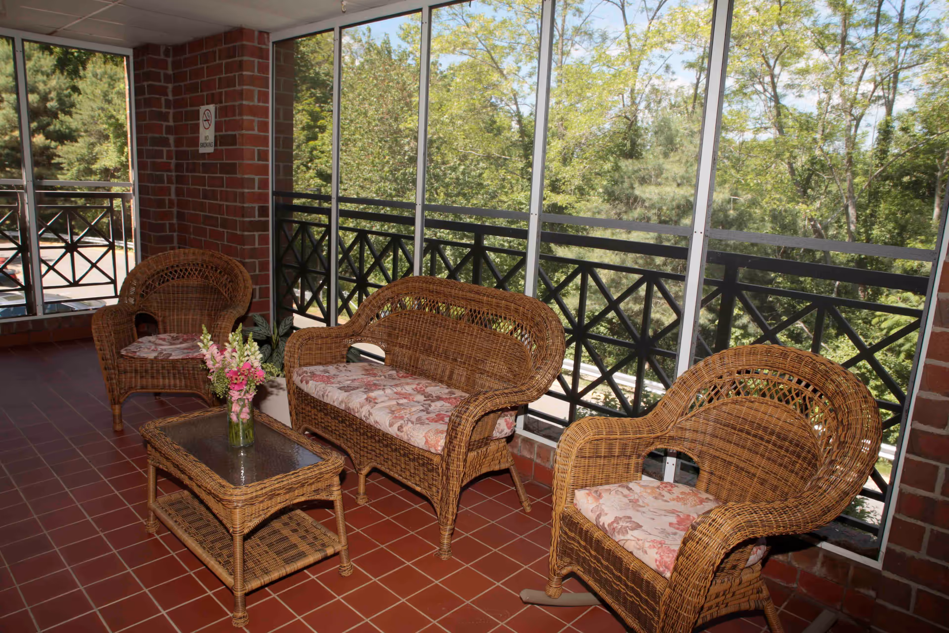 A cozy enclosed porch area with wicker furniture including a loveseat, two armchairs with floral cushions, and a glass-top wicker coffee table with a vase of flowers. The porch has large windows with black railing and a view of green trees outside. The floor is tiled with reddish-brown tiles and the walls are brick.