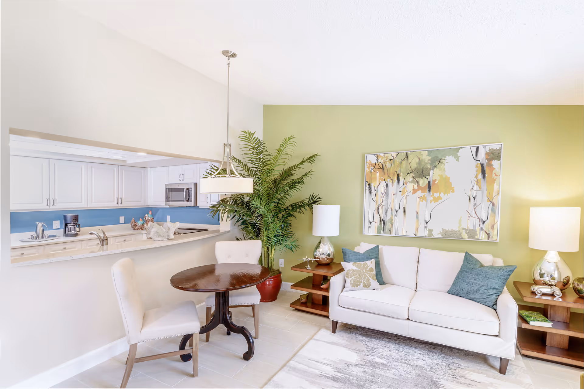 A bright and cozy living area with a white sofa adorned with blue and patterned pillows, two wooden side tables with silver lamps, a large green potted plant, and a round wooden dining table with two upholstered chairs. The background features a green accent wall with a framed abstract painting of trees and a pass-through window to a white kitchen with blue backsplash.