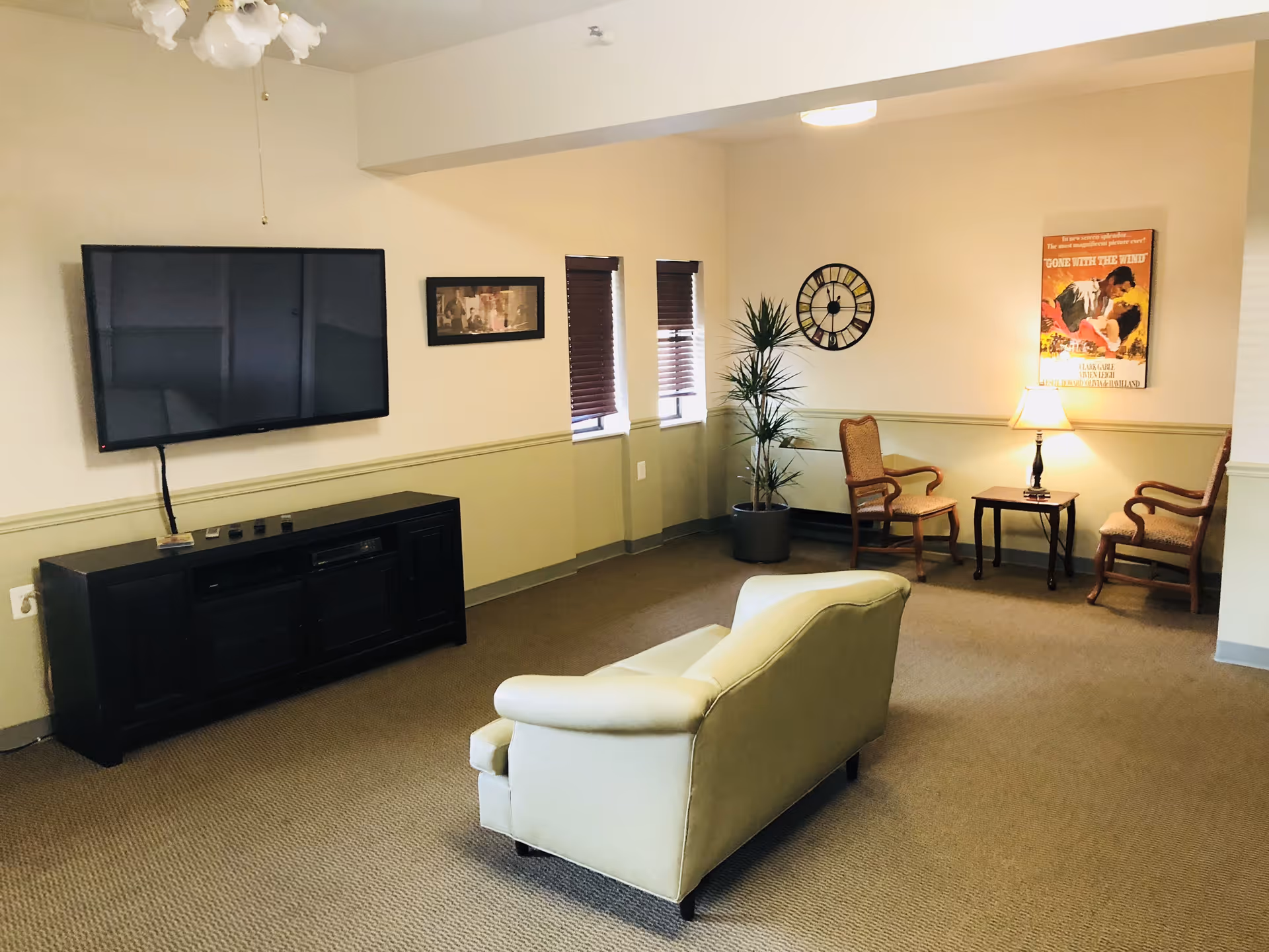 A cozy living room area with a beige loveseat facing a wall-mounted flat-screen TV above a black media console. Two small windows with brown blinds are on the wall to the right of the TV. There is a potted plant, a wall clock, and a small seating area with two wooden armchairs and a side table with a lamp. A vintage-style 'Gone with the Wind' movie poster is hung on the wall above the seating area.
