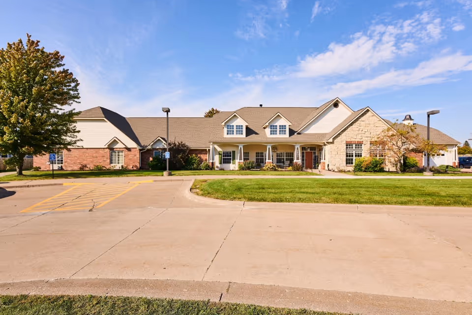 Front exterior of a single-story senior living building with a porch, landscaped lawn, and parking area under a blue sky.