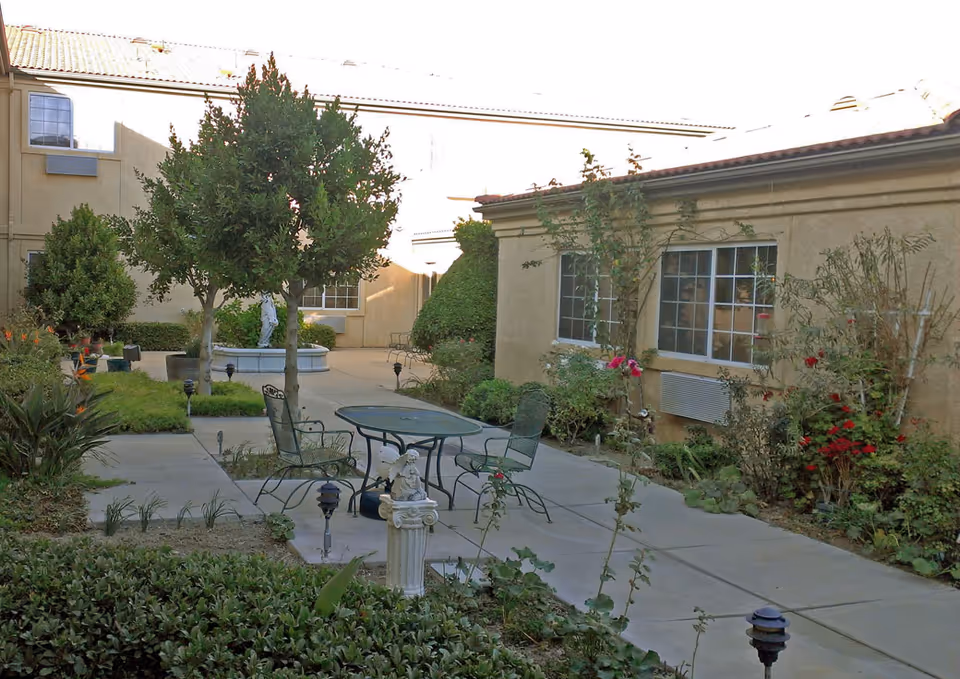 Sunlit courtyard with a patio table and chairs, a small fountain, trees, and surrounding building facades.