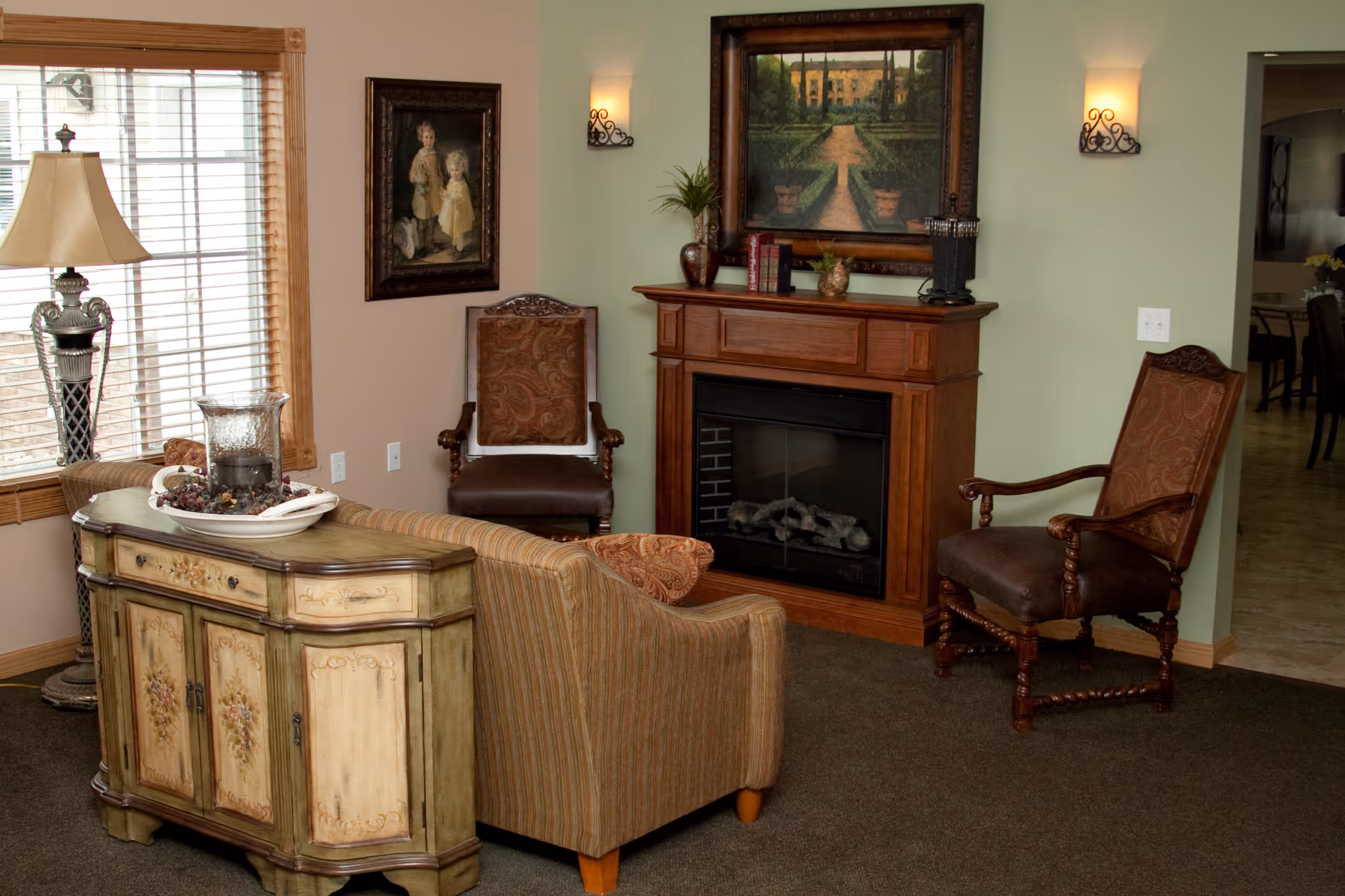 A cozy living room area with a striped sofa, two ornate wooden armchairs, a decorative wooden cabinet, and a fireplace with a framed painting above it. The room has soft lighting from wall sconces and a large window with wooden blinds.