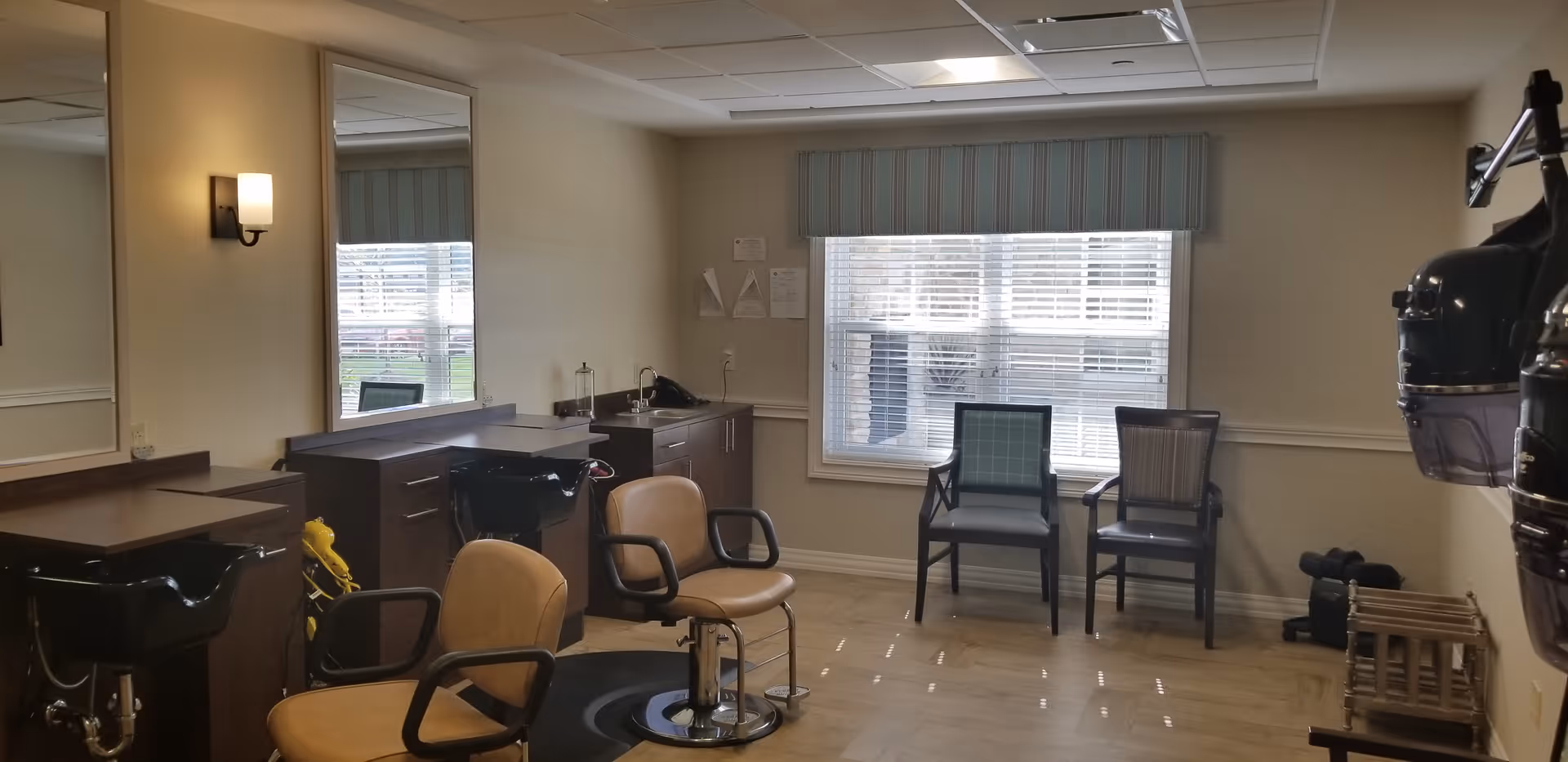 Salon-style room with styling chairs, shampoo sinks, mirrors, a hooded hair dryer, and two chairs by a large window.