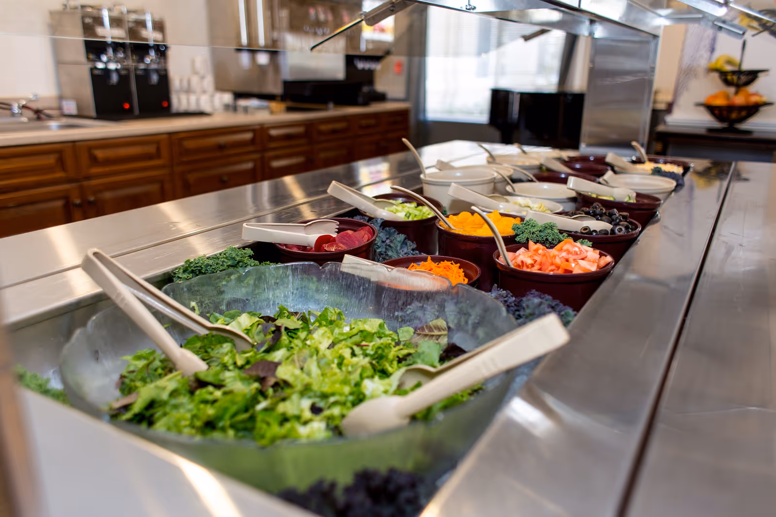 A salad bar with various fresh vegetables and toppings in bowls, including lettuce, shredded carrots, tomatoes, olives, and other salad ingredients, set on a stainless steel counter in a kitchen or dining area.